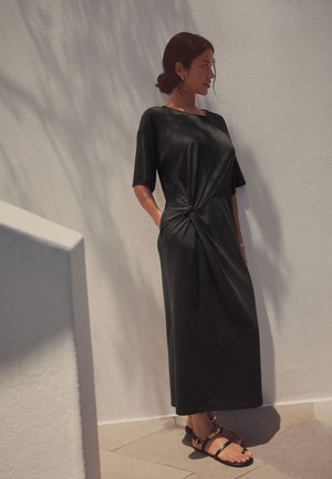 Femme aux cheveux bruns attachés en chignon, portant une robe midi noire à manches courtes avec un nœud devant, debout contre un mur clair texturé, chaussée de sandales noires.