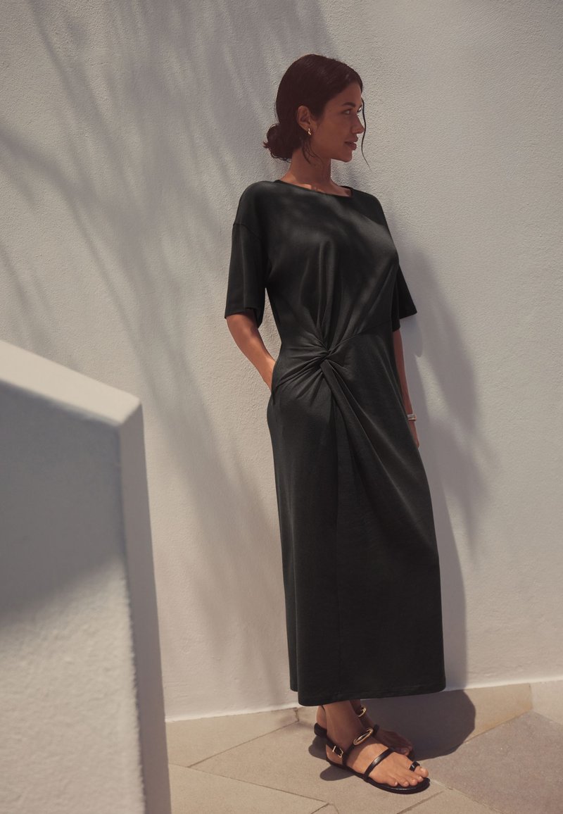 Mujer con cabello oscuro recogido en un moño, vestida con un vestido midi negro de manga corta con un nudo delantero, de pie contra una pared clara texturizada, calzando sandalias negras.