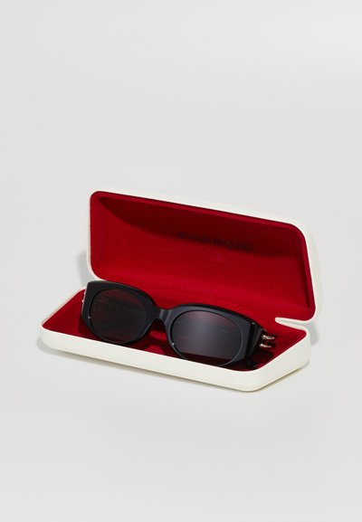 Occhiali da sole neri con lenti scure in un astuccio color crema foderato in velluto rosso. L'astuccio ha una forma rettangolare rigida.