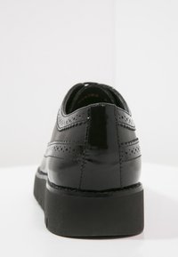 Chaussures en cuir noir à finition brillante, présentant des détails perforés au talon et une semelle épaisse en caoutchouc noir.