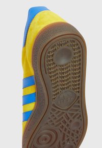 Zapatilla de ante amarilla con rayas azules. Cuenta con una suela de goma marrón texturizada y un logo redondo en el talón. Diseño aerodinámico.
