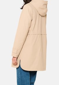 Beżowa kurtka o średniej długości z kapturem, zapięciem na napy i bocznymi rozcięciami. Wykonana z gładkiego, lekkiego materiału. Noszona na niebieskie dżinsy.