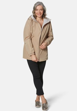 Beige geïsoleerde jas met een witte fleece-gevoerde kraag, ritssluiting en elastische manchetten. Draagt het met zwarte broeken en gestreepte instappers.