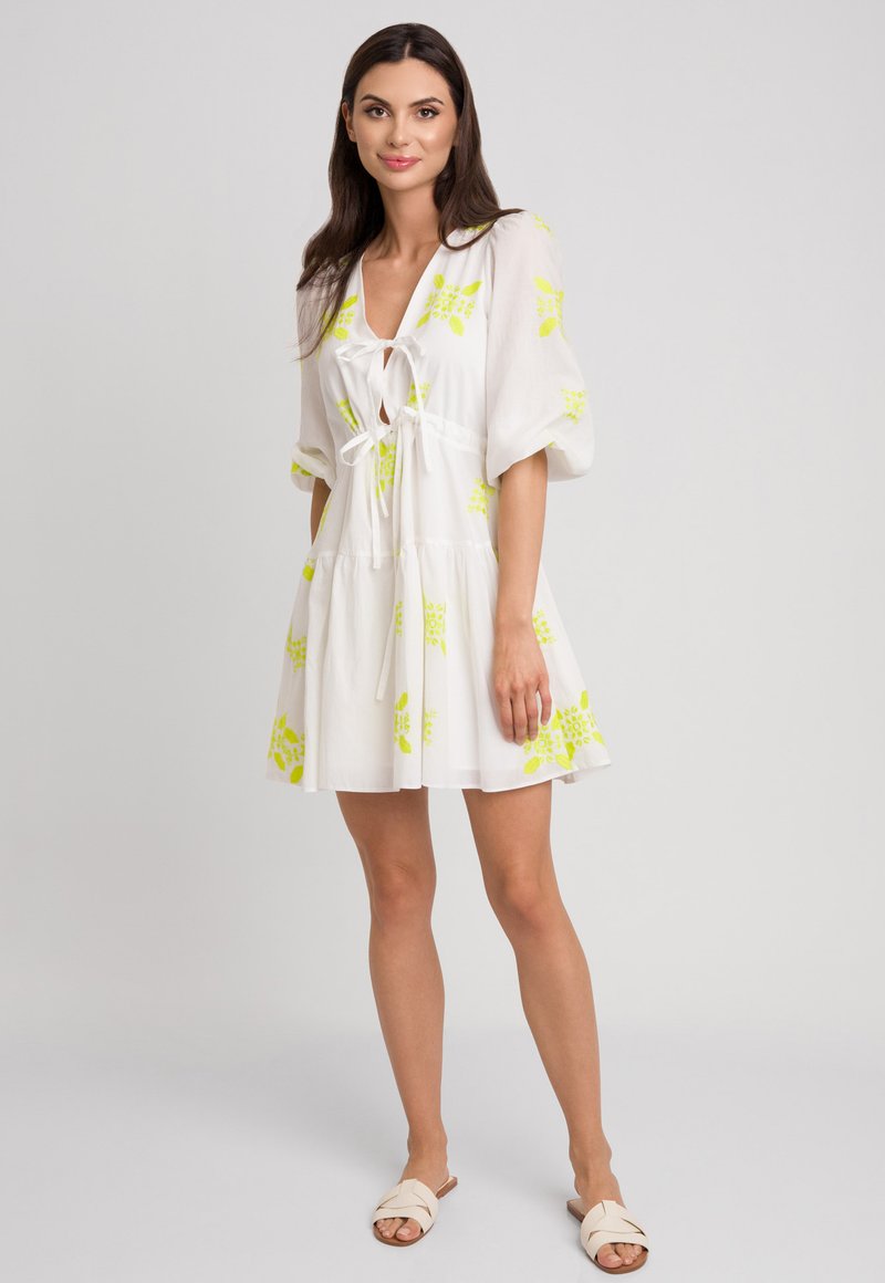 Biała krótka sukienka z bufkami, zdobiona jasnosłonecznymi wzorami kwiatowymi i szczegółem na przodzie do wiązania. Lekka tkanina, luźny krój.