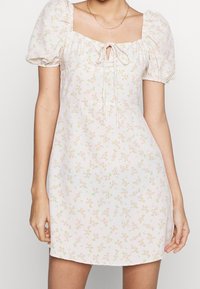 Robe mini florale en tissu rose clair avec des manches bouffantes, décolleté carré et lien à l'avant. Présente de petits motifs floraux verts et roses.