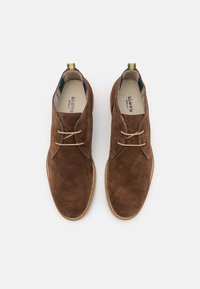 Ruske nubuk-nahka nilkkurit, joissa on vaaleanbeige nauhat, pyöreä kärki ja kontrastivärinen raidallinen yksityiskohta kantapäässä. Tiheä, teksturoitu materiaali.