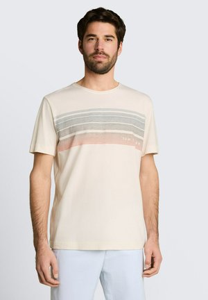 Brodaty mężczyzna ubrany w beżową koszulkę z poziomymi szarymi i różowymi paskami na klatce piersiowej, w połączeniu z jasnymi spodniami.