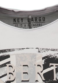 Primer plano de un cuello de camiseta blanca que muestra la etiqueta "KEY LARGO" y un diseño abstracto en blanco y negro con letras grandes, parcialmente visibles.