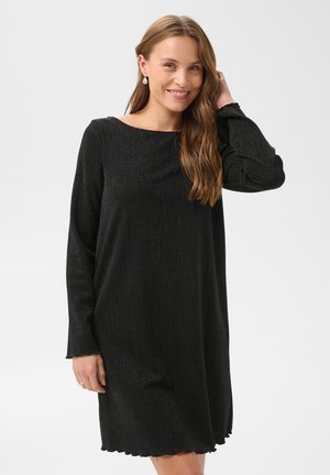 Femme aux longs cheveux bruns portant des boucles d'oreilles en perles et une robe noire à manches longues, souriante avec une main touchant ses cheveux.