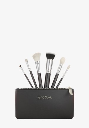 Six pinceaux de maquillage avec manches noirs et viroles argentées dans une trousse noire zippée partiellement ouverte étiquetée ZOEVA.