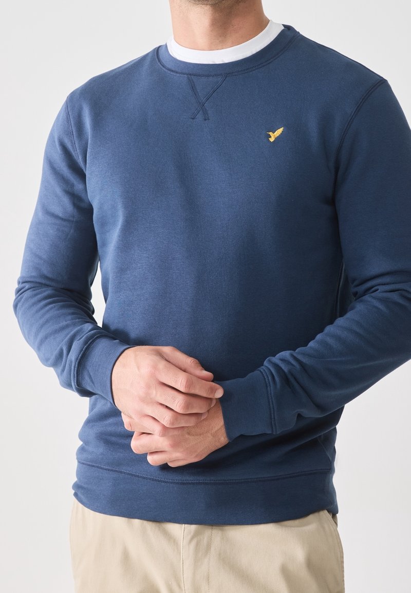 Homme portant un sweat-shirt bleu marine à col rond avec un petit logo oiseau jaune sur la poitrine et un pantalon beige, mains jointes devant.