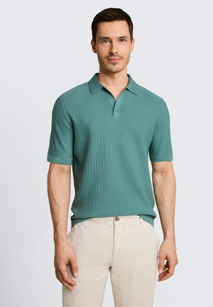 Mann trägt ein kurzärmeliges, geripptes Poloshirt in Petrol und hellbeige Hose, steht vor einem schlichten hellen Hintergrund.