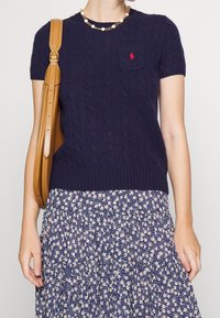 Osoba v námořnickém svetr s krátkým rukávem pleteným do copánků, květinové sukni, s perlovým náhrdelníkem a nese hnědou koženou kabelku přes rameno.