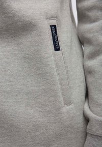 Sudadera gris claro hecha de una tela suave, con un bolsillo lateral y una etiqueta con texto azul. Textura suave y un diseño simple y moderno.