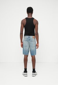 Débardeur noir, short en denim bleu clair avec une finition délavée et un patch en cuir rectangulaire à l'arrière, porté avec des chaussettes noires et des chaussures blanches.