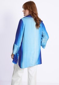 Blouse fluide à dégradé de bleus, avec des manches longues, un col, des fentes sur les côtés et un tissu léger et doux.