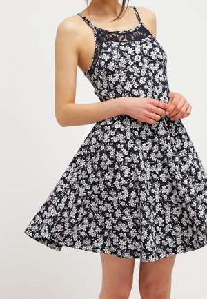 Zwarte mouwloze jurk met wit bloemenpatroon en kantdetail bij de halslijn, gedragen door een persoon die voor een effen achtergrond staat.