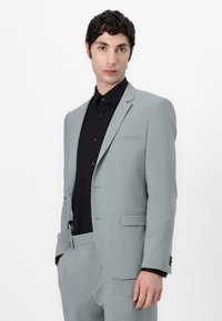 Lichtgrijs op maat gemaakt kostuum met een gladde textuur en notched revers, gedragen over een zwart shirt. Heeft een enkele borstzak en subtiele stikseldetails.