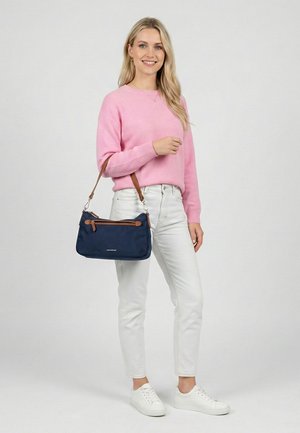 Smilende kvinde iført lyserød sweater, hvide jeans, hvide sneakers, der holder en navyblå taske med brune stropper mod en ensfarvet baggrund.