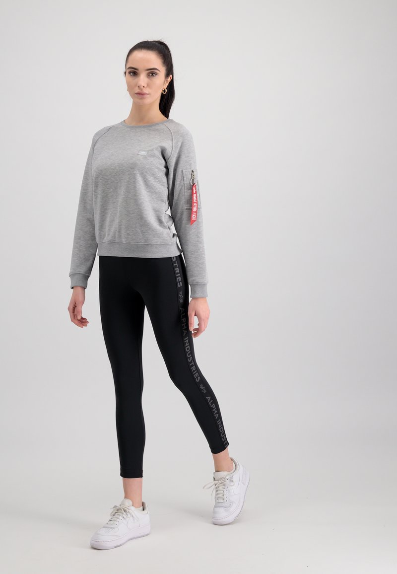 Sivý sveter s vreckom na zips na rukáve, skombinovaný s čiernymi legínami s potlačou loga. Model nosí biele tenisky.