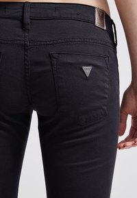 Jeans de mezclilla negros con un acento de logo metálico en forma de triángulo en el bolsillo trasero, con un diseño ajustado y una textura suave.