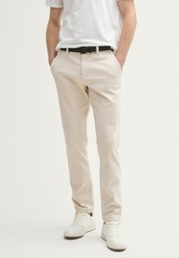 Homme portant un chino beige avec une ceinture noire, des baskets blanches et une chemise blanche à manches courtes, se tenant les mains dans les poches contre un fond uni.