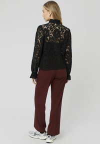 Blouse en dentelle noire à manches longues, col haut, motif floral, associée à un pantalon bordeaux et des baskets blanches. Détail en dentelle texturé visible.