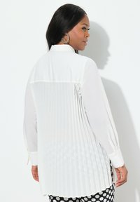 Witte geplooide blouse met lange mouwen, kraag en knoopmanchetten. Beschikt over een soepele textuur, plooien aan de achterkant en valt onder de taille.