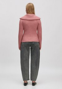 Frau mit langen blonden Haaren, die einen rosa gerippten Pullover mit weitem Kragen, lockere graue Jeans und schwarze Schuhe trägt, steht mit dem Rücken zur Kamera.