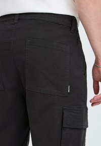 Black cargo pants with textured fabric, featuring large side pockets and a small label tag on the rear. Simple, functional design.