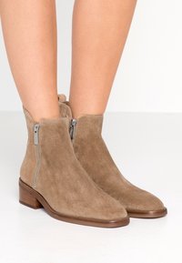 Zamszowe botki do kostki w kolorze taupe z zaostrzonym noskiem i dwoma srebrnymi suwakami po zewnętrznej stronie. Masywne drewniane obcasy zapewniają stabilność.