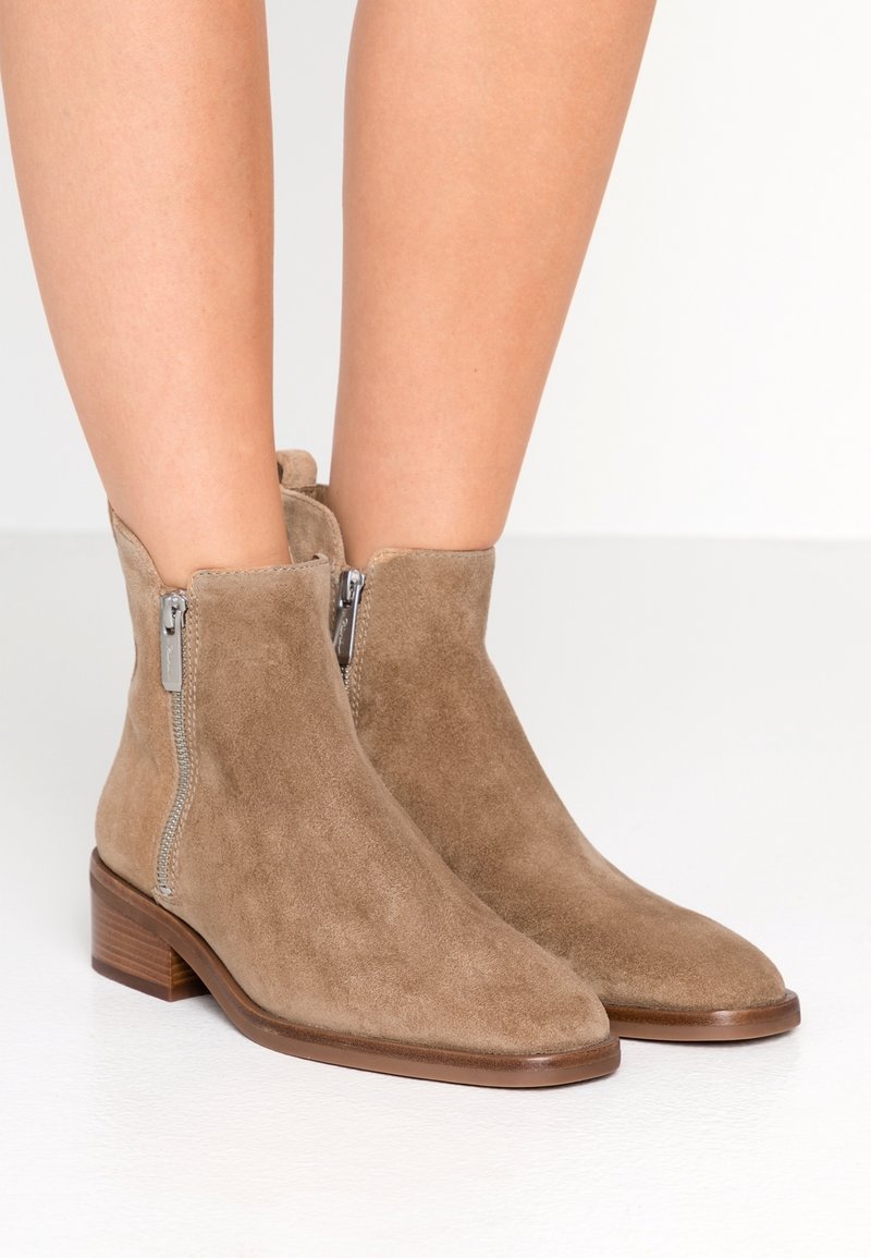 Zamszowe botki do kostki w kolorze taupe z zaostrzonym noskiem i dwoma srebrnymi suwakami po zewnętrznej stronie. Masywne drewniane obcasy zapewniają stabilność.