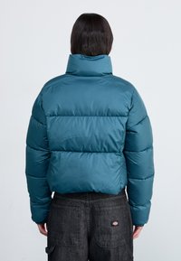 Teal dunsjakke med høy krage, quiltet mønster og blank finish. Brukt med svarte denimbukser med baklommer.