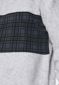 Grå fleecetröja med en marinblå rutigt ullpanel över bröstet. Tyget är mjukt med en borstad yta; subtila gula och vita accenter i mönstret.