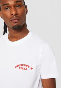 Weißes T-Shirt aus Baumwolle mit einem roten aufgestickten Logo, das "GIUSEPPE’S PIZZA" auf der linken Brust zeigt. Einfaches Design mit Rundhalsausschnitt.