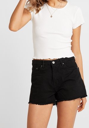 Femme portant un crop top blanc côtelé à manches courtes et un short en jean noir taille haute avec des ourlets effilochés et des colliers superposés.