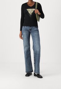 Kvinna i svart långärmad tröja med guld- och vit "GUESS" logotyp, blå raka jeans, svarta loafers och bärande på en grön axelväska.