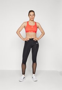 Red sports bra with white logo and matching band; black leggings with mesh panels; white sneakers with black stripes.