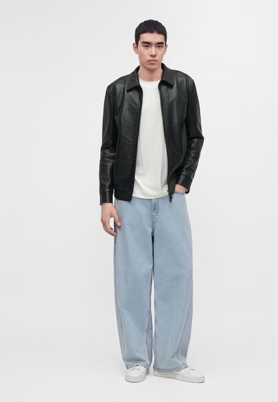 Young man wearing black leather jacket, white t-shirt, wide light blue jeans, and white sneakers against plain background.