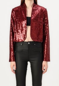 Red sequined cropped jacket with long sleeves, featuring a shawl collar. Paired with tight black pants, showcasing a sleek and shiny texture.