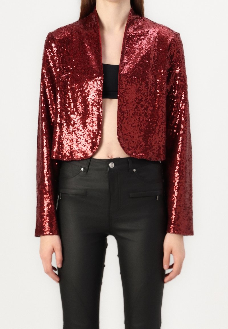 Red sequined cropped jacket with long sleeves, featuring a shawl collar. Paired with tight black pants, showcasing a sleek and shiny texture.