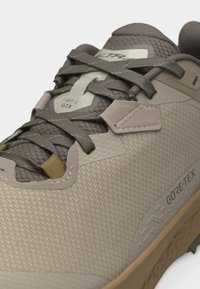 Κλείσιμο πλάνου ενός πεζοπορικού παπουτσιού σε μπεζ και καφέ αποχρώσεις με υφασμάτινη υφή, ετικέτα Gore-Tex και δεμένα διαγώνια κορδόνια πάνω σε επενδεδυμένη γλώσσα με την ένδειξη "Timp 6 GTX".