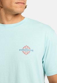 Vaaleansininen puuvillapaita, jossa on pieni monivärinen Billabong-logo ja koristeellinen kuvio rinnassa, pyöreä pääntie ja lyhyet hihat.