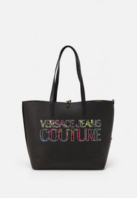 Black tote bag with dual handles and "Versace Jeans Couture" logo in multicolor floral pattern on front.
