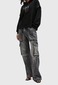 Černá mikina s kapucí a potiskem na přední straně, doplněná o šedé cargo kalhoty s nášivkovými kapsami a opotřebovanými detaily. Sandály s dřevěným podpatkem.