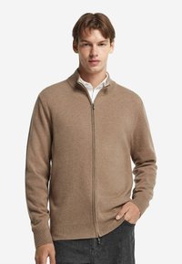 Beige gestrickter Reißverschluss-Pullover mit hohem Kragen, gerippten Bündchen und Saum; zeichnet sich durch eine glatte Textur und subtile Farbvariationen aus.