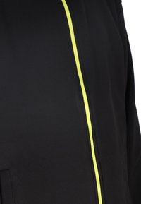 Schwarze Jacke mit einem auffälligen vertikalen Reißverschluss in neon-gelb und einem glatten, strukturierten Stoff. Keine weiteren Details sichtbar.