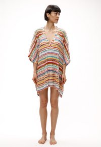 Γυναίκα με κοντά σκούρα μαλλιά, όρθια και ξυπόλυτη, φοράει φαρδιά πολύχρωμη φόρεμα με ζιγκ-ζαγκ σχέδιο, φαρδιά μανίκια και V-λαιμόκοψη, κοιτώντας προς τα δεξιά της.