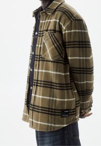 Camisa chaqueta de cuadros marrón con rayas negras, confeccionada en tela texturizada, con un bolsillo en el pecho y botones a presión. Etiqueta negra en el dobladillo.