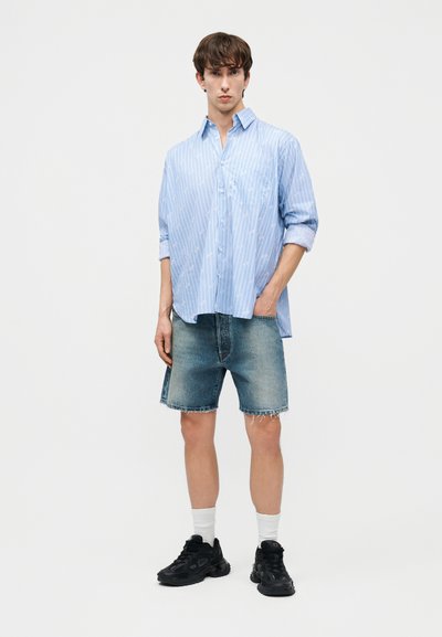 Jauns vīrietis, kurš valkā gaiši zilu svītrainu krekla pogu kreklu ar savilktām piedurknēm, džinsa šortus, baltas zeķes un melnus sporta apavus, stāvot pret vienkāršu fonu.